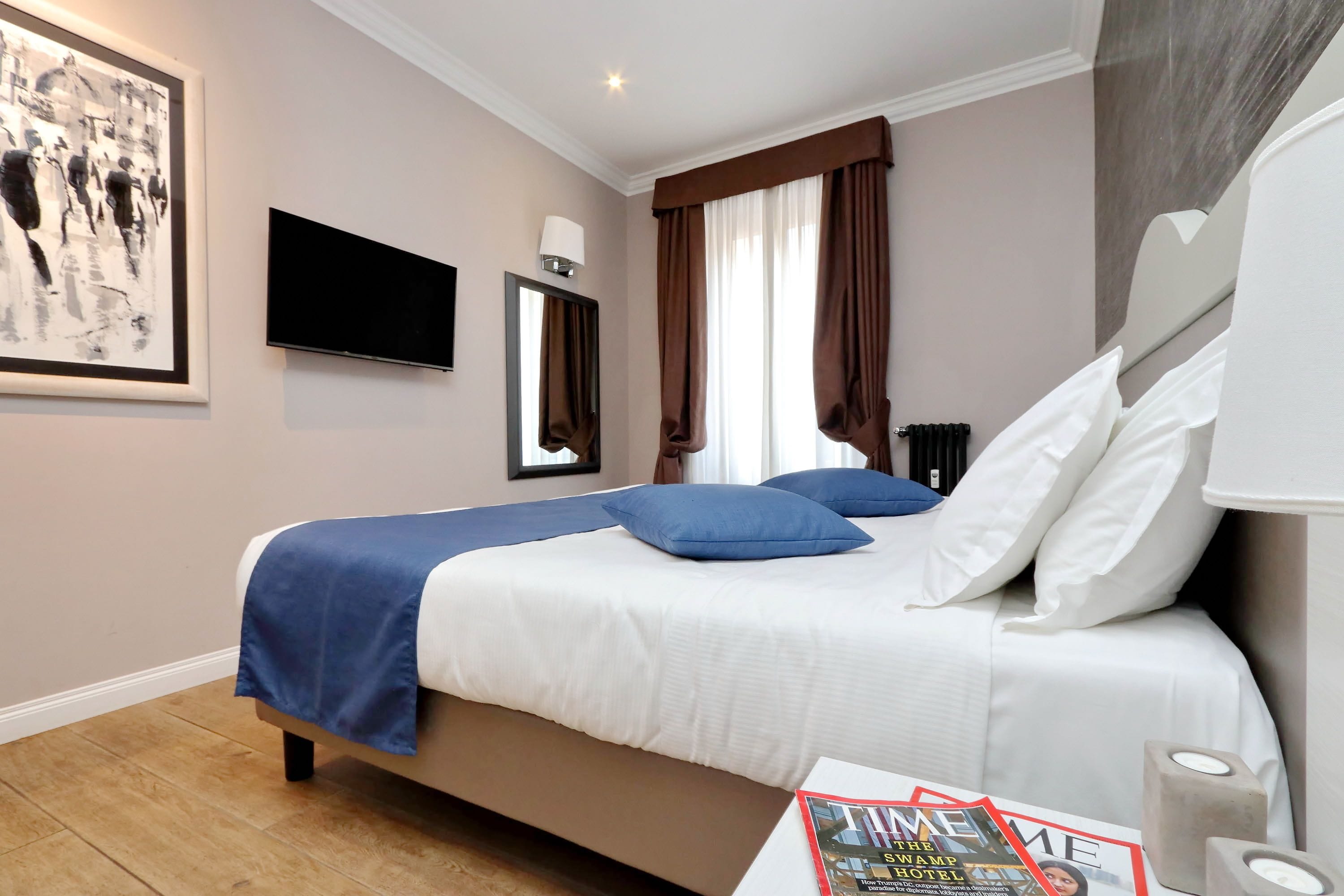 A hotel room with a bed, a flat-screen TV, a framed artwork, a window with curtains, and a magazine on the nightstand.