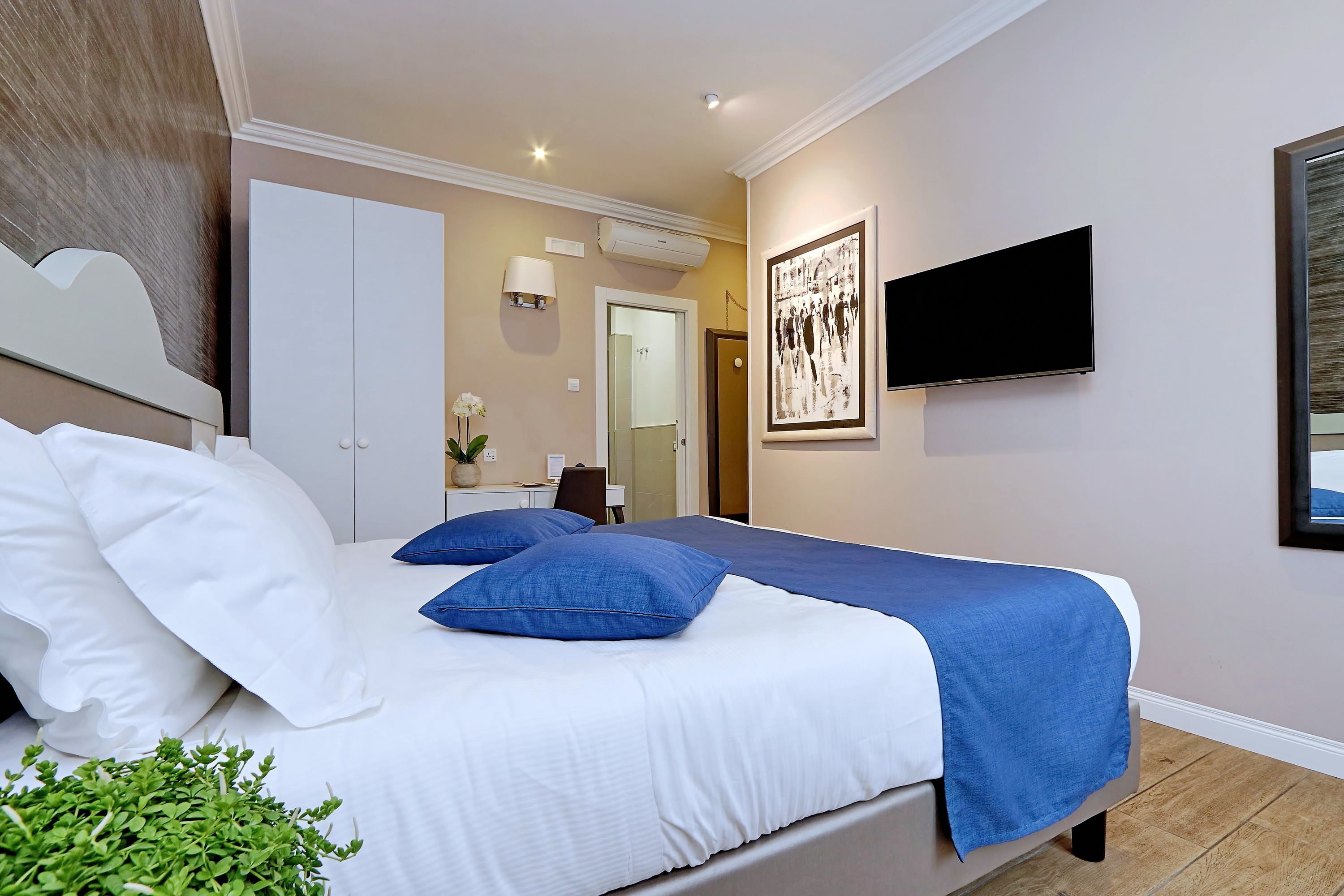 Een slaapkamer met een bed, beddengoed in wit en blauw, een televisie en een ingelijste foto aan de muur.