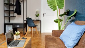 A modern living room with a brown leather sofa, a laptop on a wooden table, a black metal shelving unit with a coat hanging, a blue chair, and a large green plant. - Vico Rooms and Terrace (Rome)