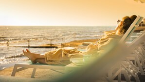 A person relaxing on a sun lounger by the beach with a book and a glass of water.