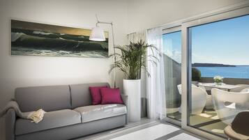 Uma sala de estar moderna com um sofá cinza, uma planta grande em vaso e vista para o mar através de uma porta de vidro.
