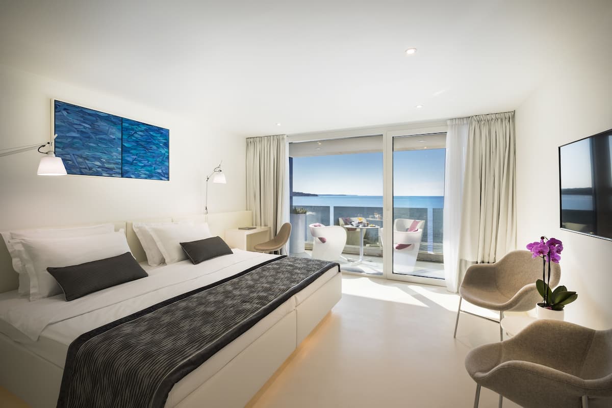 A modern bedroom with a large bed, a desk, and a balcony with a view of the sea.