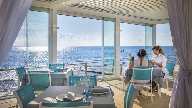 A dining area with tables and chairs, large windows with a view of the sea, and a clear sky.