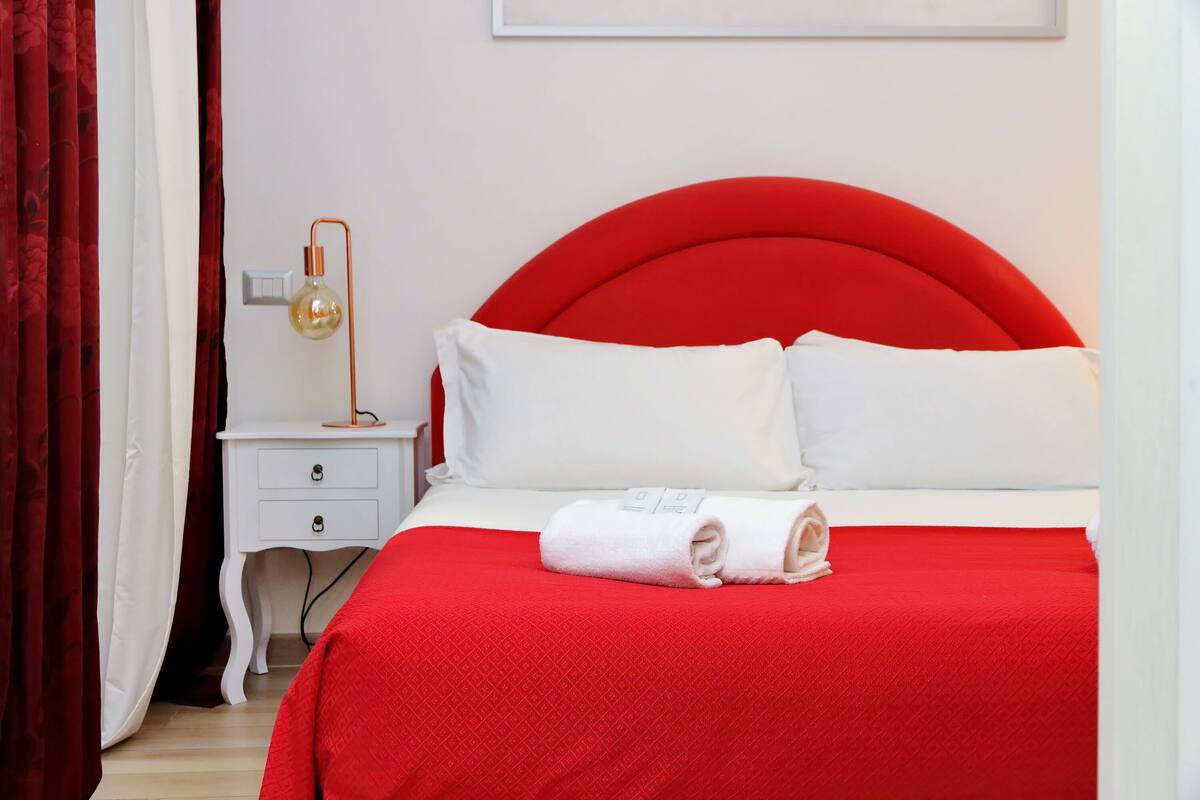 A neatly made bed with a red headboard and white bedding, a bedside lamp, and a white nightstand with drawers.