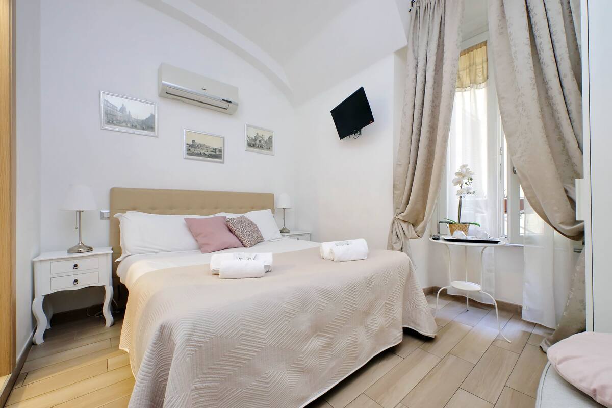 A neatly arranged bedroom with a bed, bedside tables, a flat-screen TV, and framed pictures on the wall.