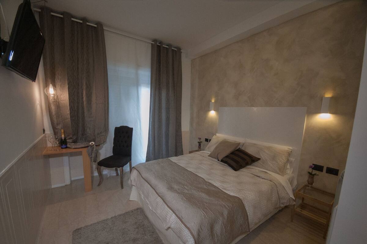 A bedroom with a large bed, a chair, a TV mounted on the wall, and a small table with a bottle and glass.
