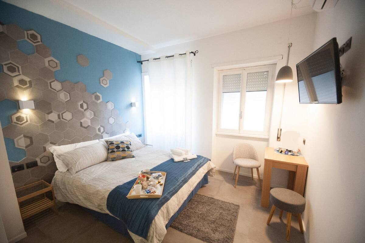 A bedroom with a bed, a desk, a chair, a TV, and a window with curtains.