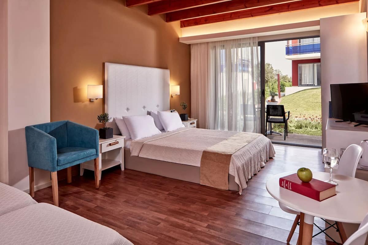 Una habitación de hotel moderna con una cama grande, un sillón azul, un escritorio con una computadora portátil, un televisor y un balcón con vistas al exterior.