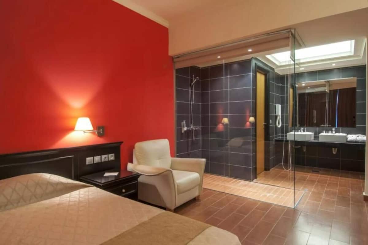 Habitación de hotel con pared de acento roja, cama con cubierta color beige, mesita de noche, sillón blanco, zona de ducha con cerramiento de vidrio y baño con un espejo grande y dos lavamanos.