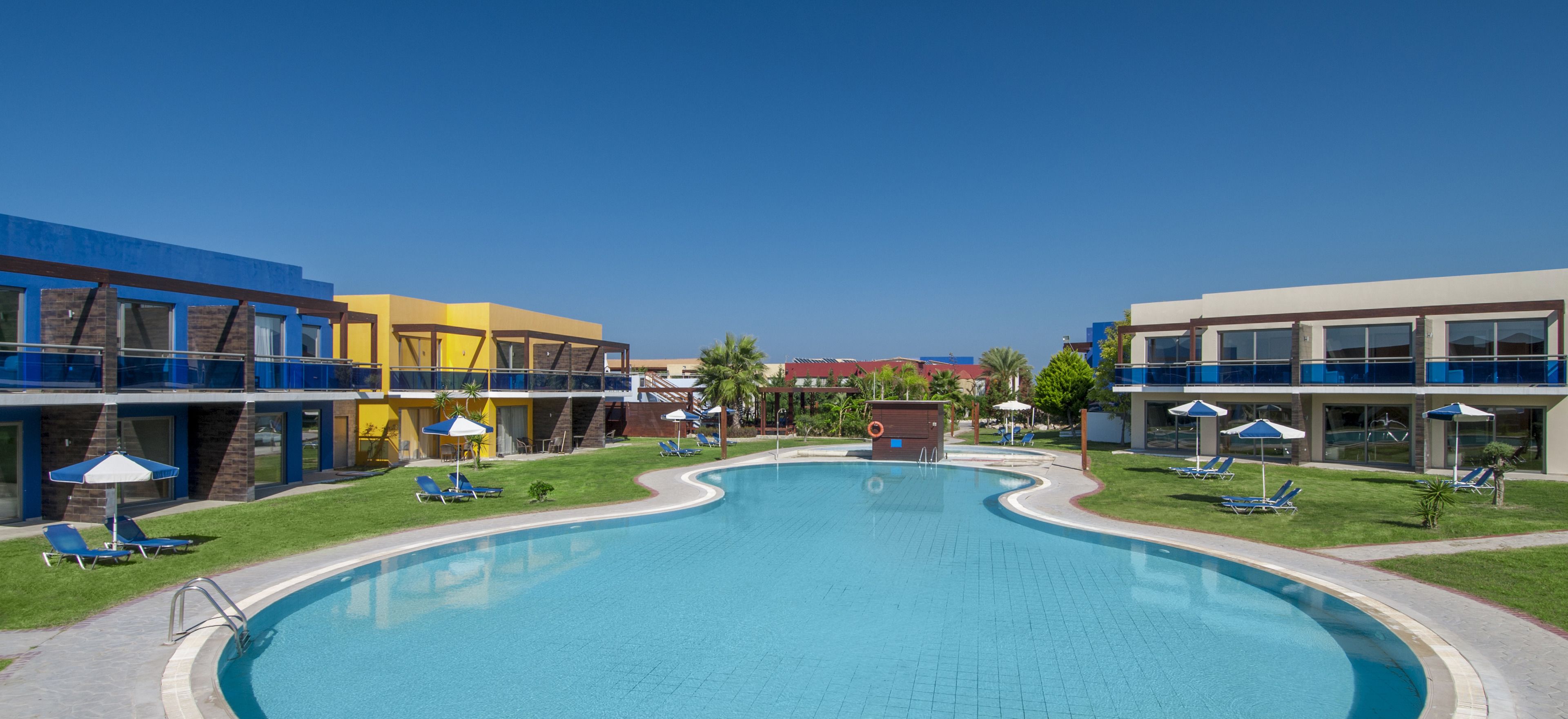 A swimming pool surrounded by modern buildings with balconies, sun loungers, and umbrellas.