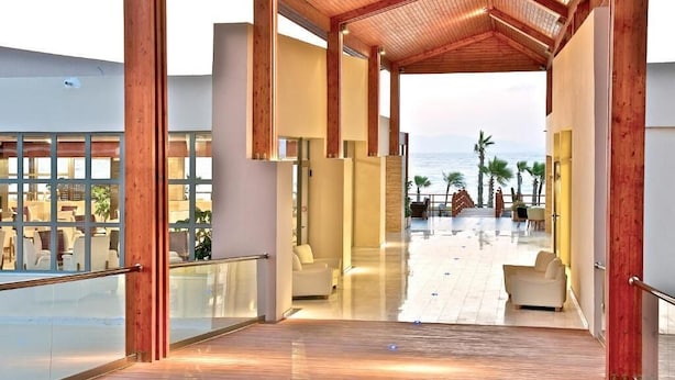 A spacious indoor area with a wooden ceiling and large windows, leading to an outdoor patio with seating and a view of the sea.