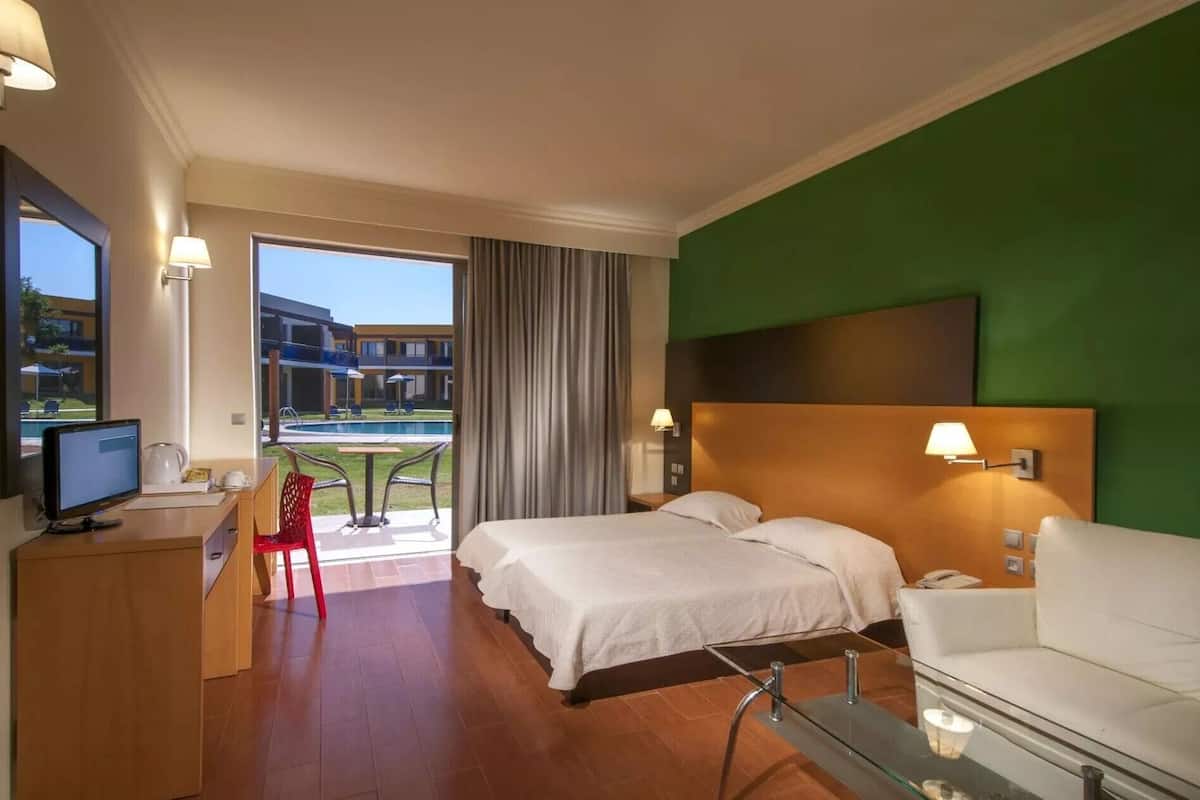 Habitación de hotel con cama, escritorio con computadora, silla, mesa de vidrio, sofá y vista a una piscina y edificios.