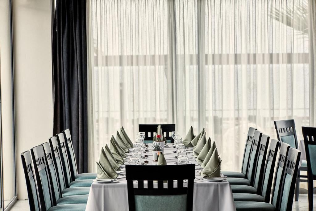 A dining room with a long table set for a meal, flanked by chairs, and large windows with sheer curtains.