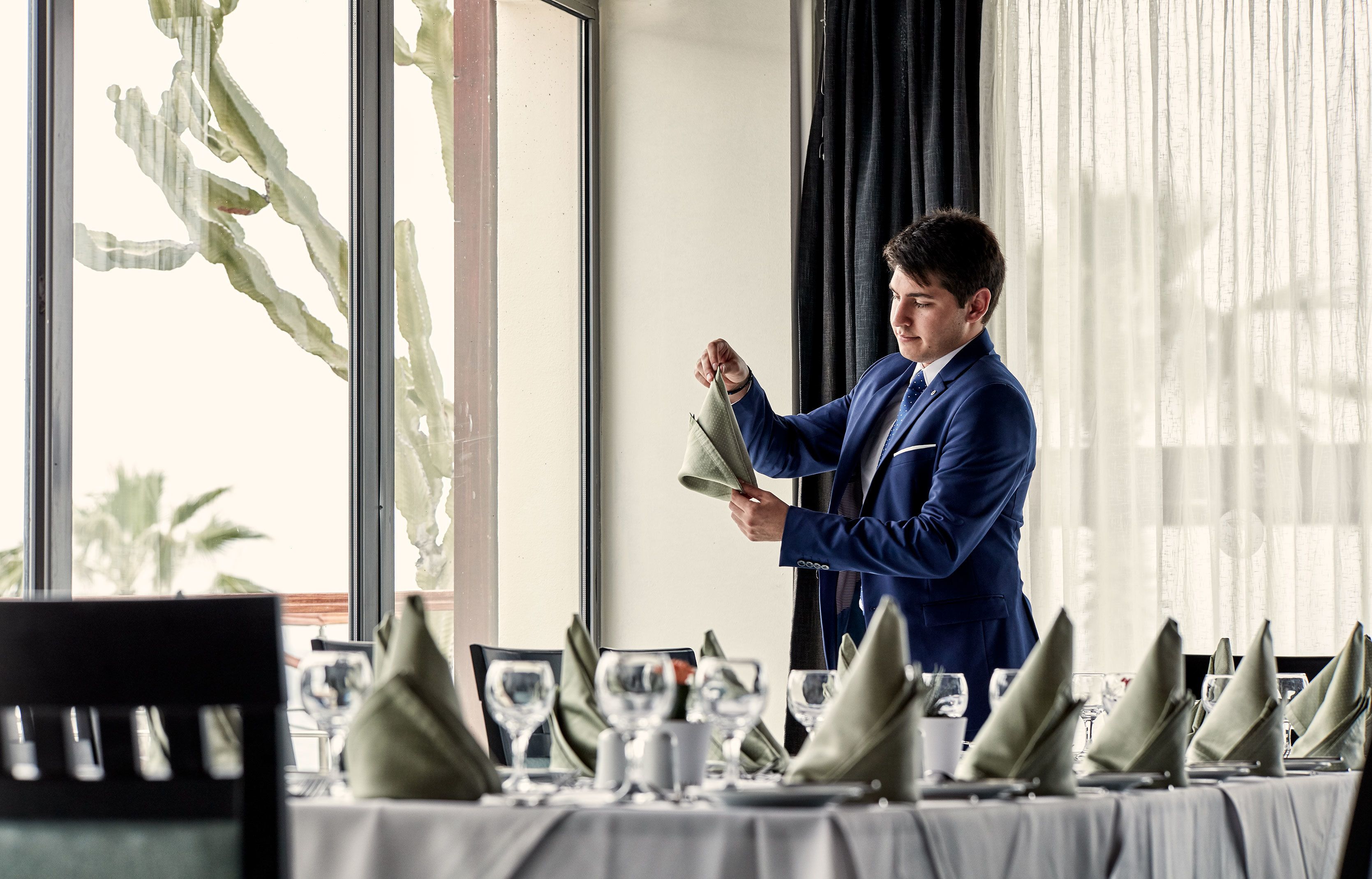 Eine Person in einem blauen Anzug bereitet einen Tisch mit grünen Servietten und Besteck in einem hellen Essbereich mit großen Fenstern vor.