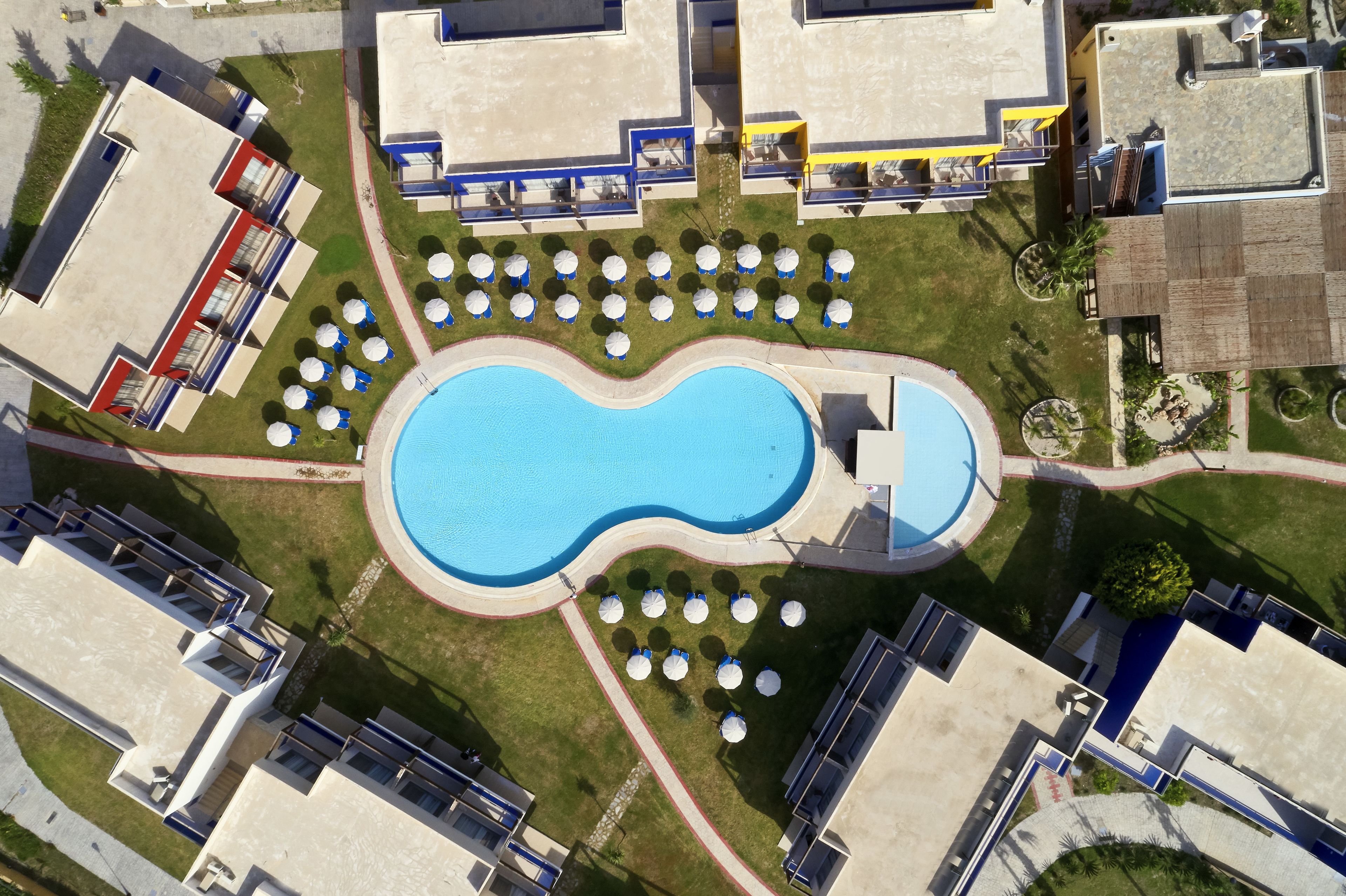 Ein Luftbild eines Hotels mit Schwimmbad, umgeben von Liegestühlen und Sonnenschirmen, verbunden mit mehreren Gebäuden mit Balkonen.