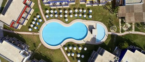 Ein Luftbild eines Hotels mit Schwimmbad, umgeben von Liegestühlen und Sonnenschirmen, verbunden mit mehreren Gebäuden mit Balkonen.