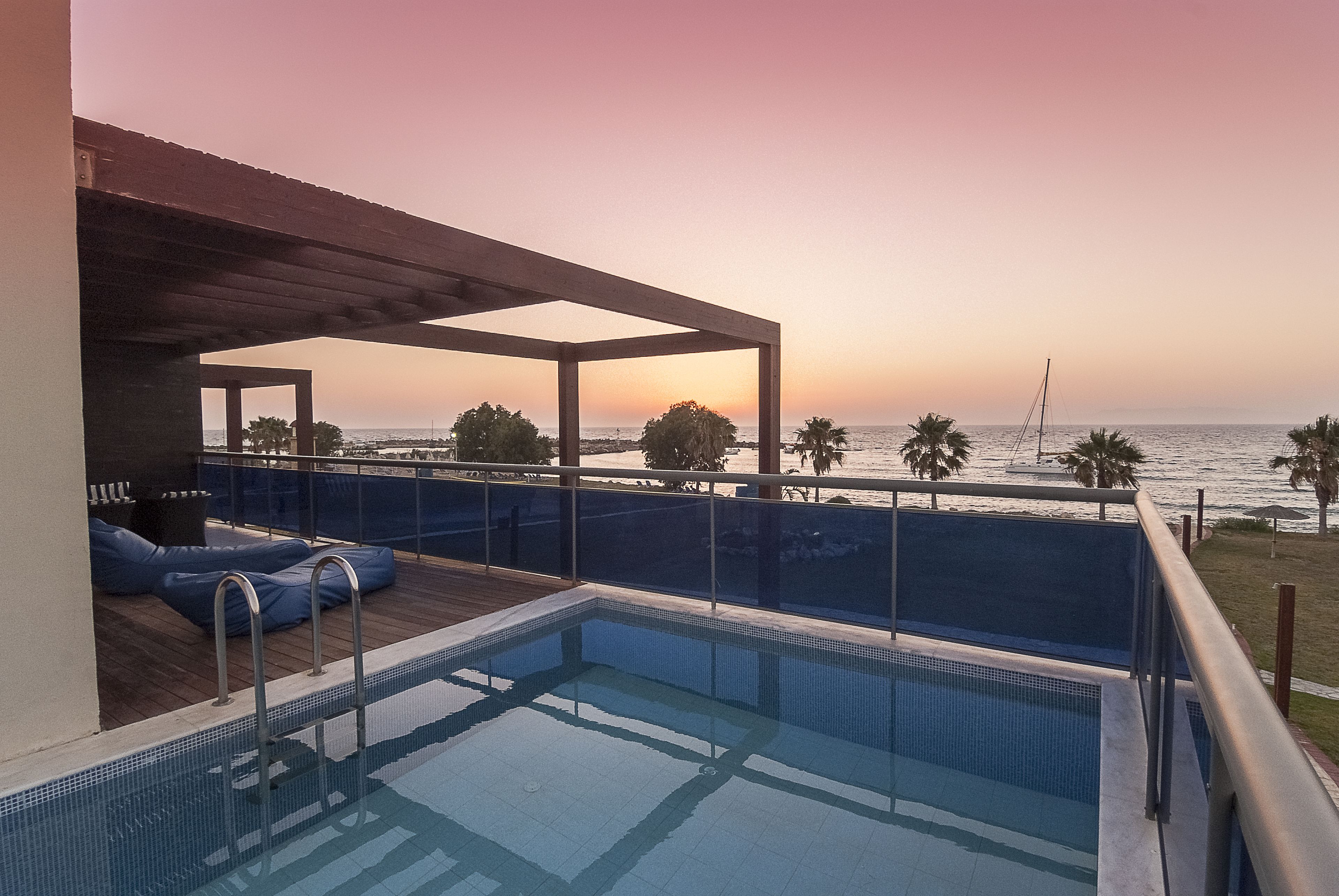 A pool area with a view of the ocean at sunset, a covered seating area, and a deck with lounge chairs.