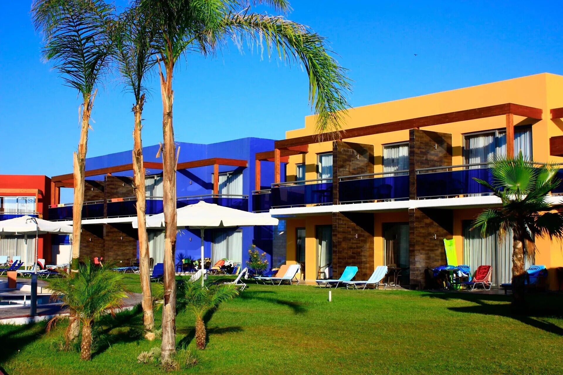 Un hotel de diseño moderno, con zona de piscina con sillones de descanso y sombrillas, y un césped cuidado con palmeras.