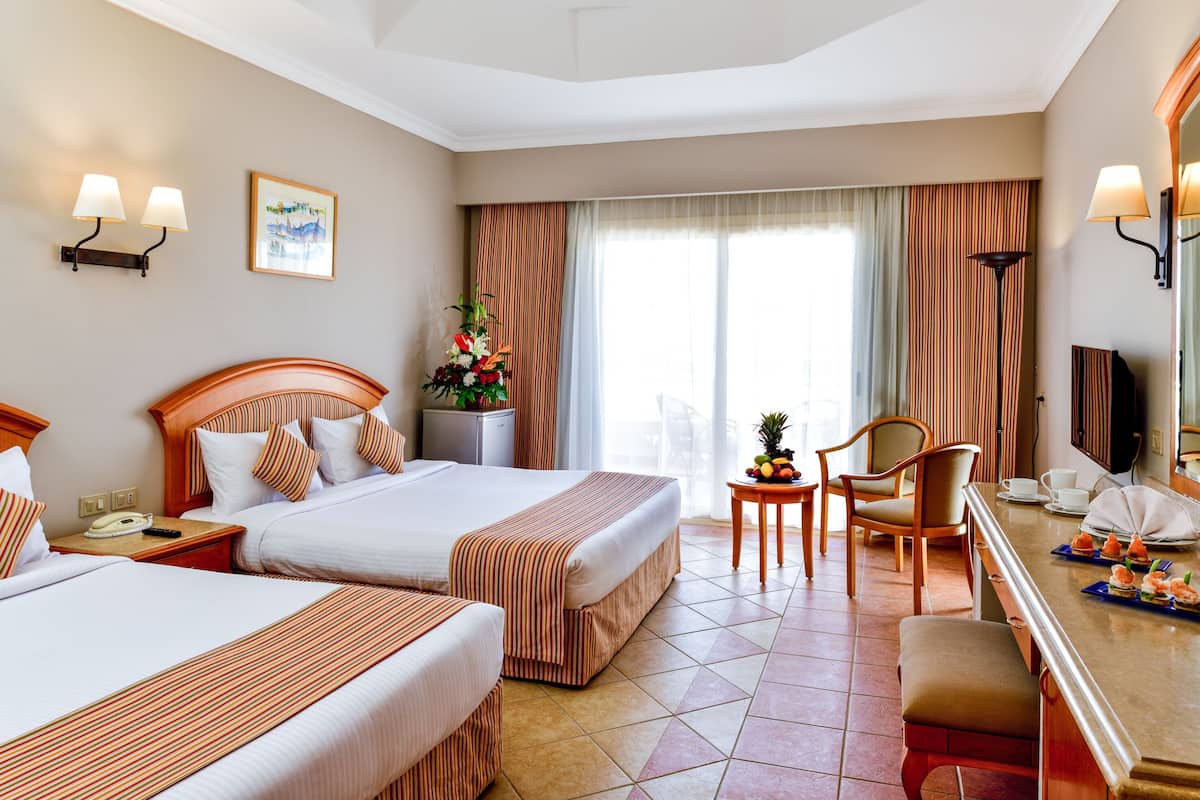A hotel room with two beds, a desk, a chair, a small table with fruit, and a large window with curtains.