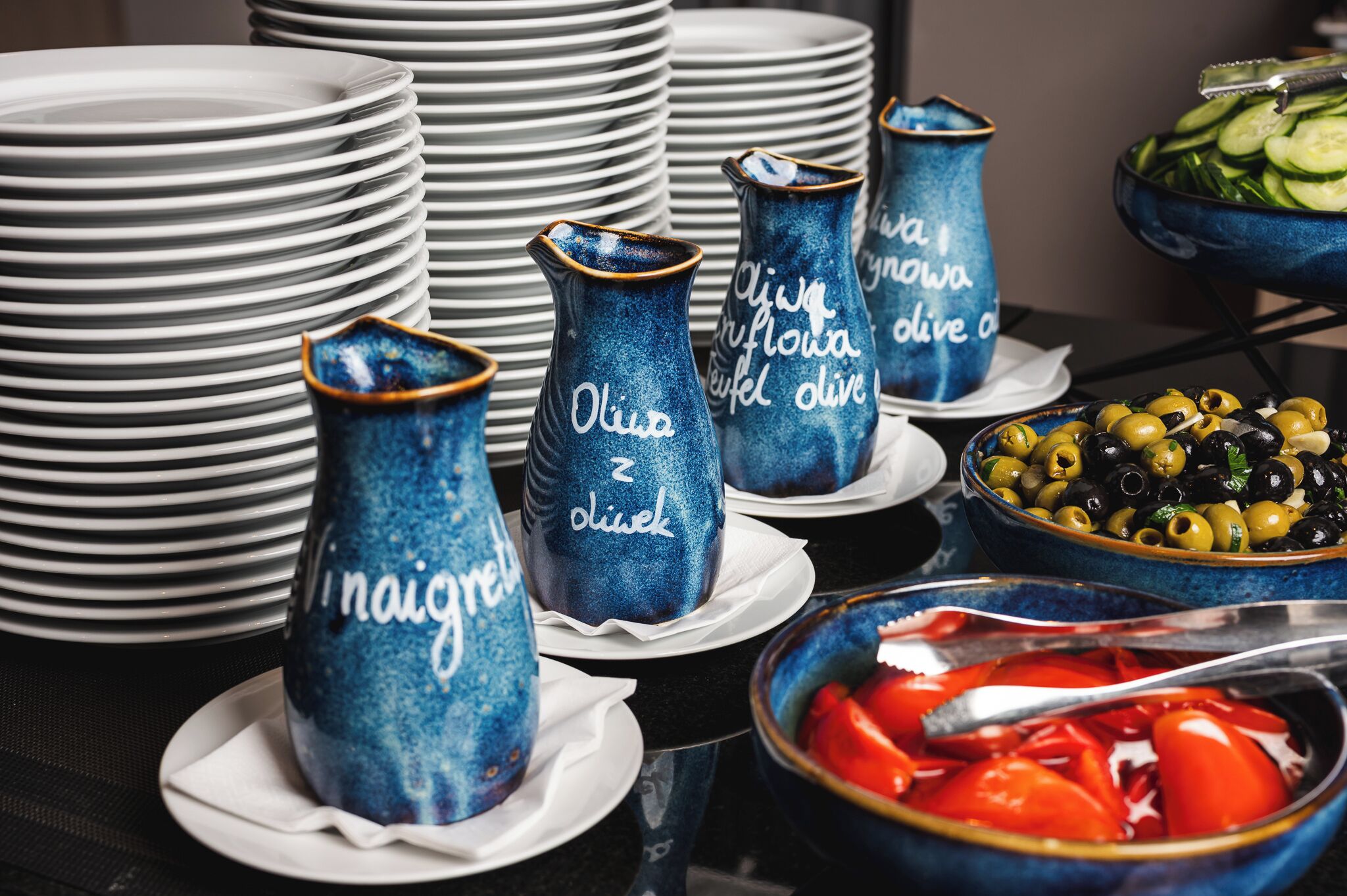 A buffet setup with blue vases labeled "Olive & Zoliek," white plates, and bowls of olives and sliced tomatoes.