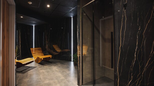 Een moderne, overdekte ruimte met een donkere marmeren wand, een glazen deur en houten lounge fauteuils.