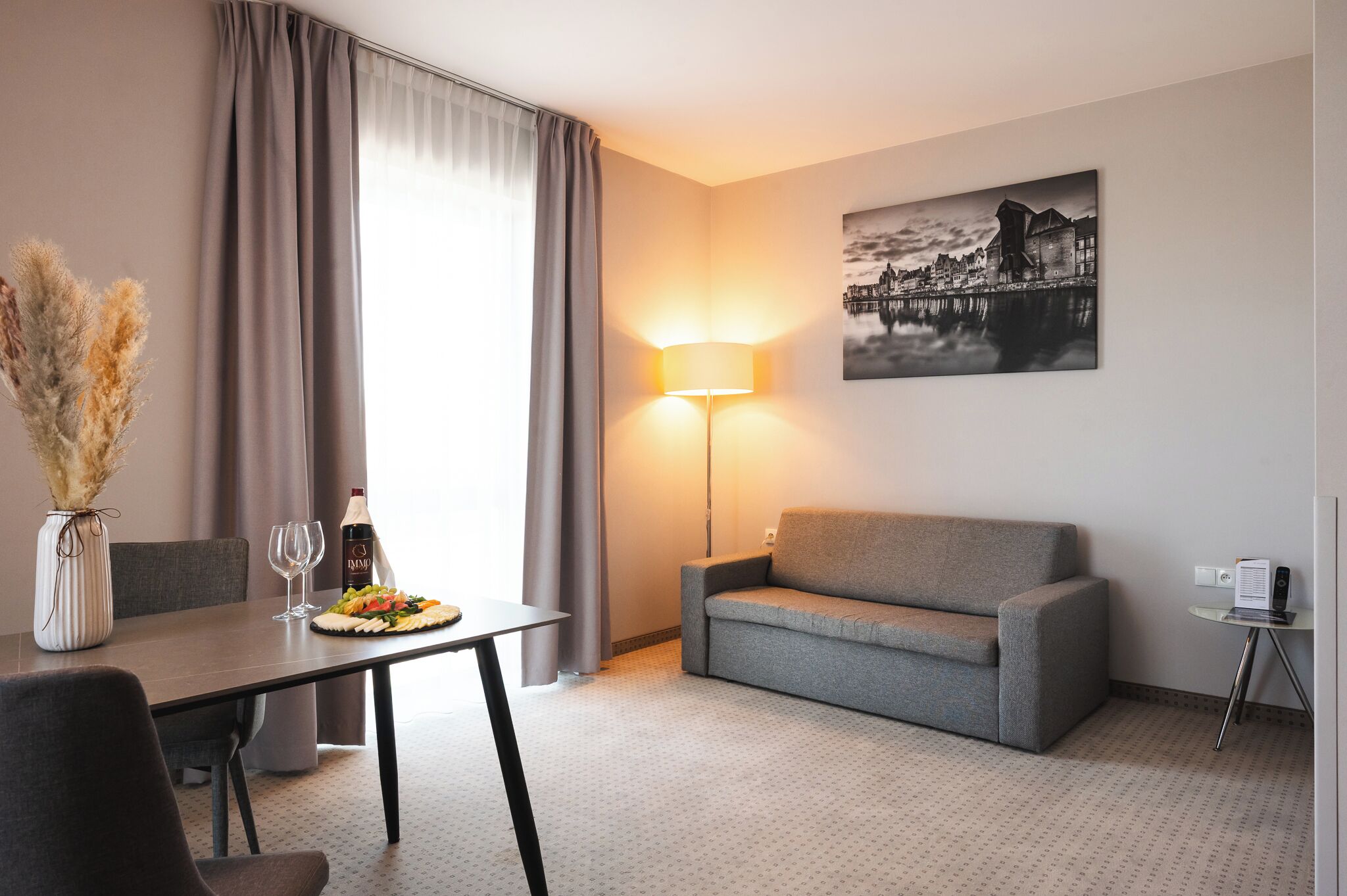Ein Hotelzimmer mit einem Esstisch, gedeckt mit einer Flasche, Gläsern und einem Teller mit Essen. Ein graues Sofa und eine Lampe auf einem Beistelltisch sind ebenfalls vorhanden. An der Wand hängt ein Gemälde, das ein Gebäude zeigt.