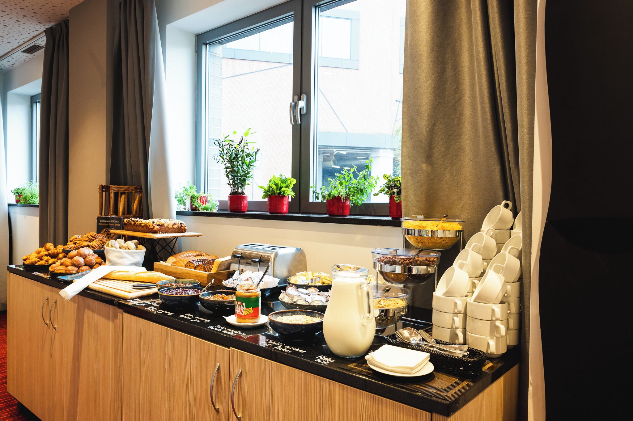 Un bufé de desayuno con diversas opciones de comida, una máquina de café y plantas en macetas junto a la ventana.