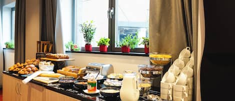 Un bufé de desayuno con diversas opciones de comida, una máquina de café y plantas en macetas junto a la ventana.
