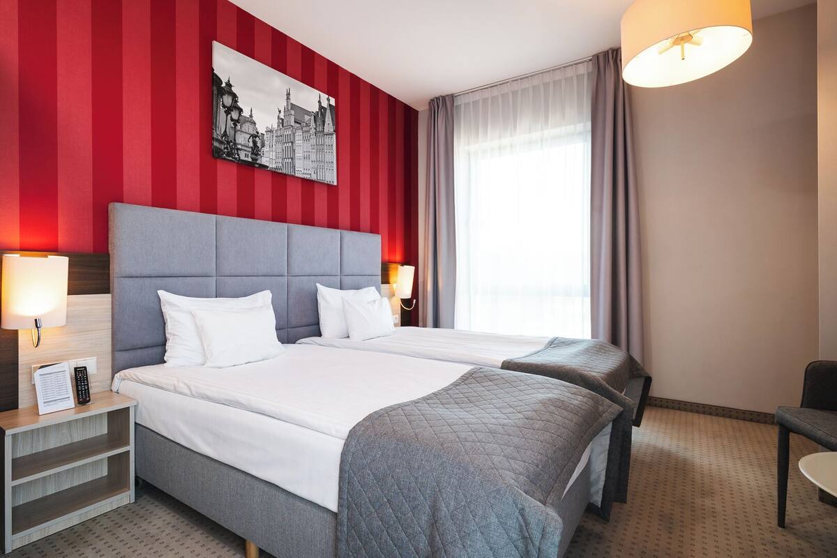 Ein Hotelzimmer mit zwei Betten, einer roten Wand mit Streifen, einem Stadtbildgemälde und einem großen Fenster mit Vorhängen.