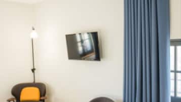 A hotel room with two armchairs, a small table, a floor lamp, a TV on the wall, and a large window with curtains.