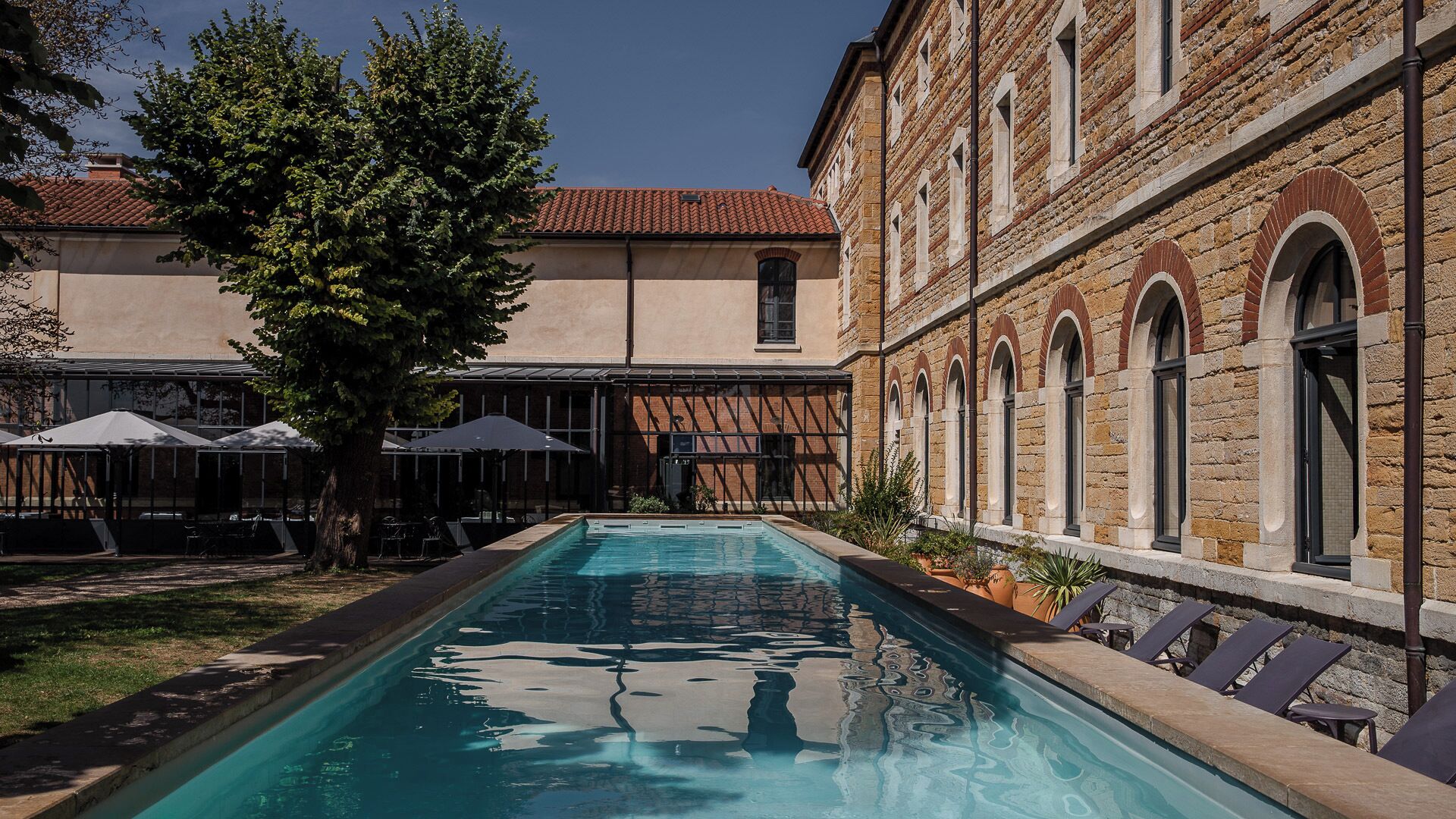 A swimming pool with lounge chairs, a covered seating area, and a building with arched windows.