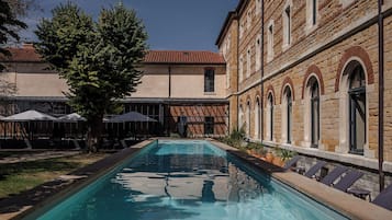 Une piscine avec des chaises longues, un espace de détente couvert et un bâtiment doté de fenêtres en arc de cercle.