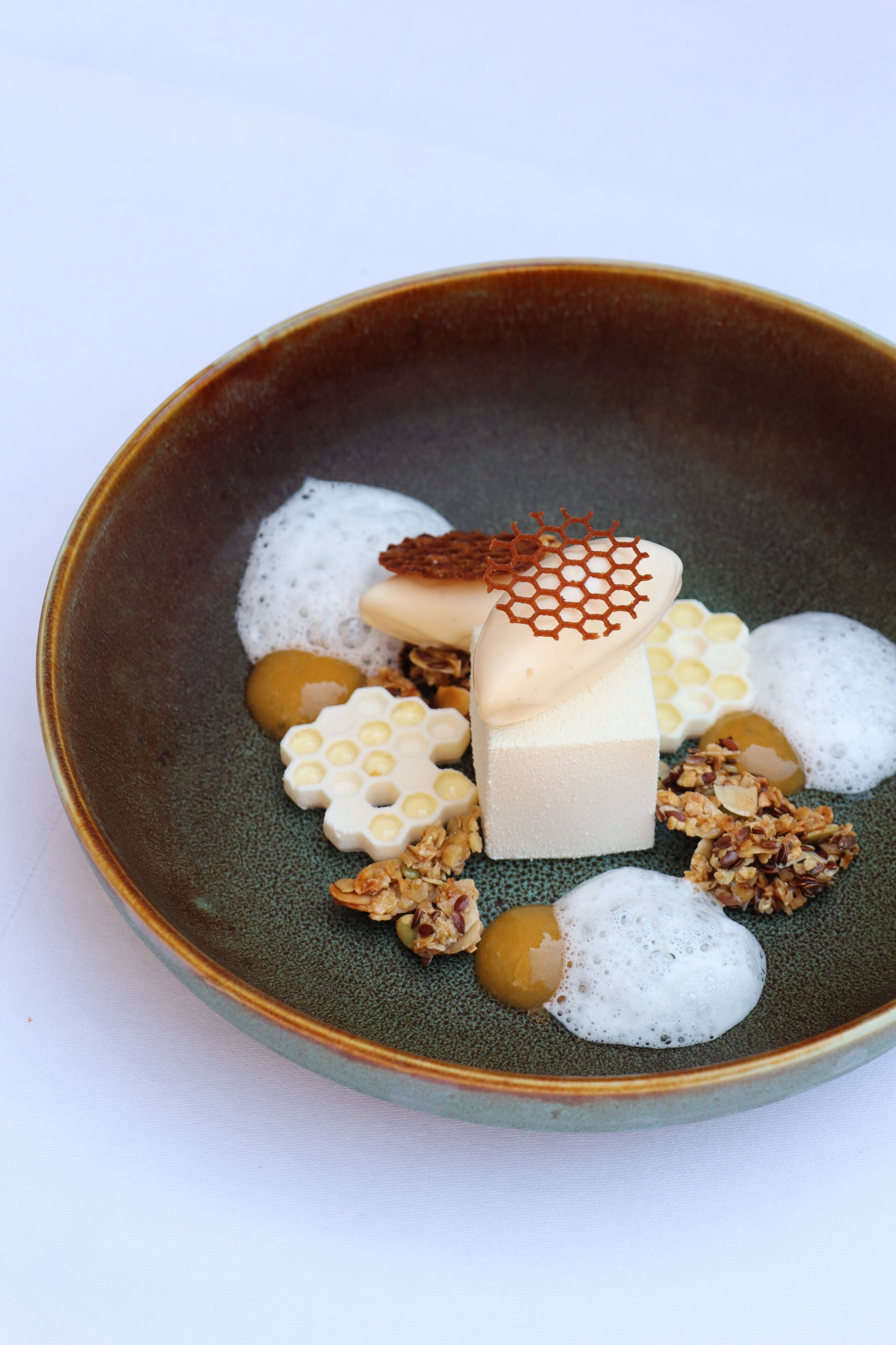 A ceramic bowl containing a dessert with honeycomb, a cube of white substance, and granola.