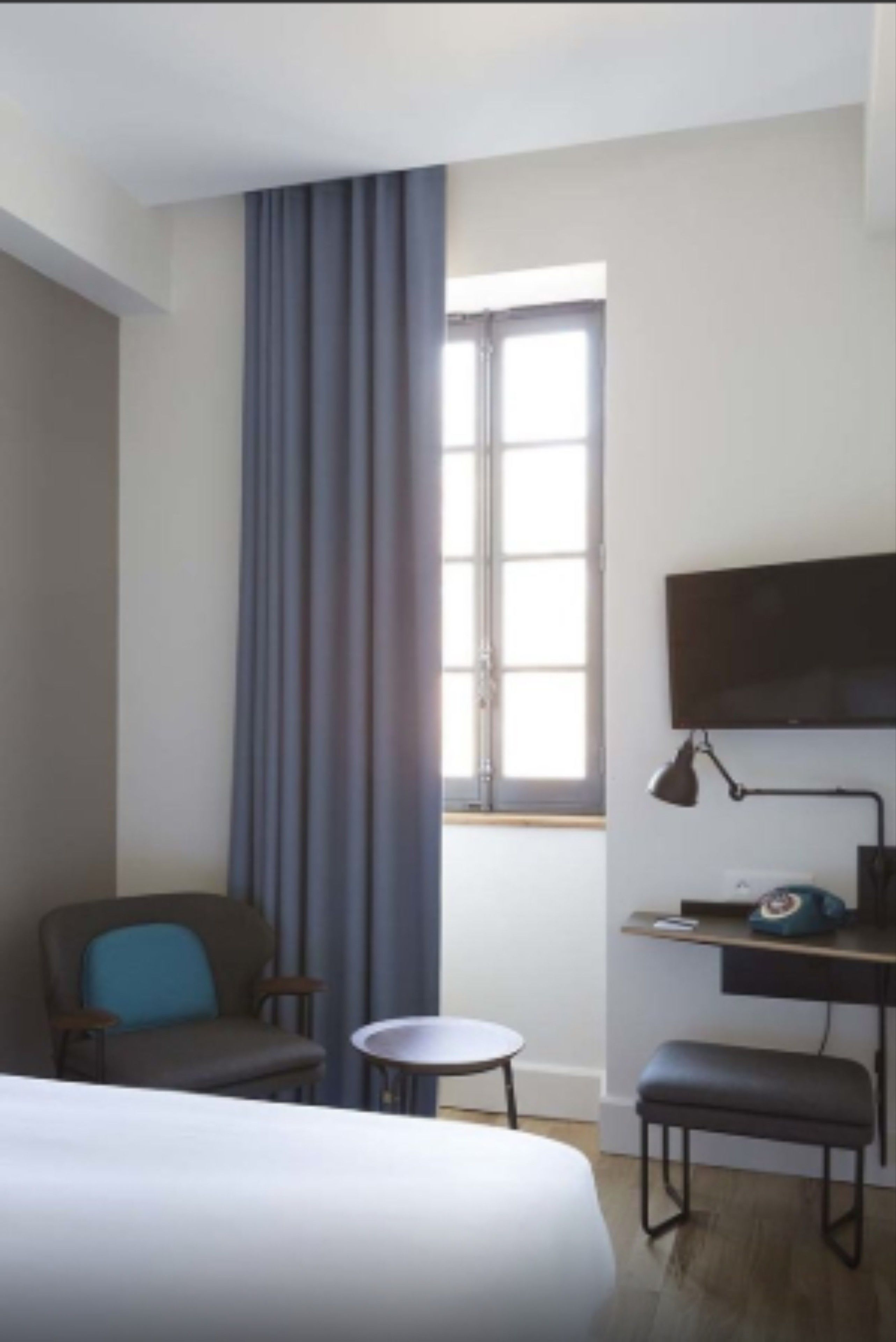 A hotel room with a bed, a desk with a lamp, a chair, a small table, and a TV.