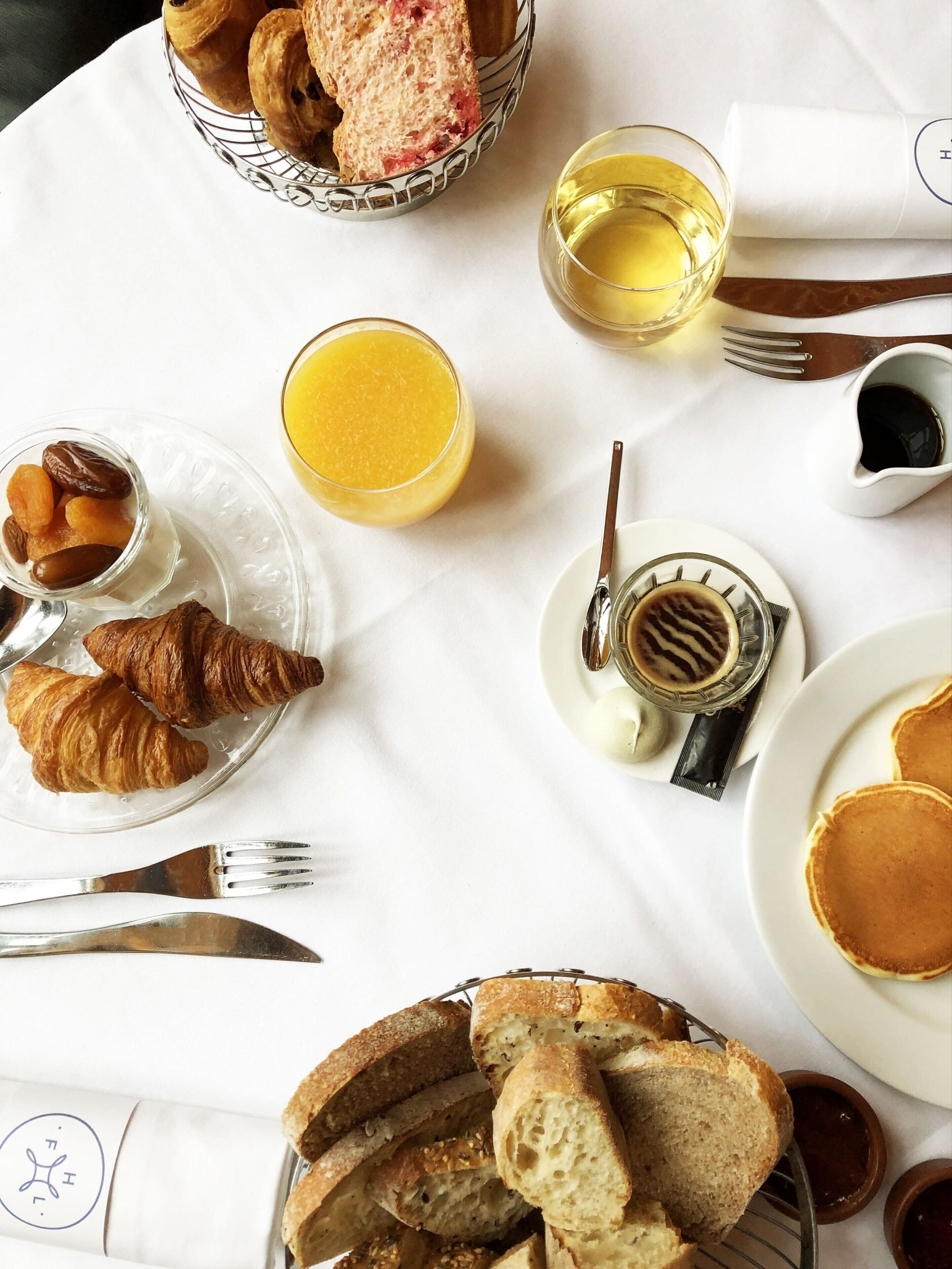Una mesa de desayuno con cruasanes, pan, tortitas, un vaso de zumo de naranja y una taza de café.