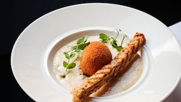 Un piatto bianco con un dessert composto da un boccone rotondo impanato, una fetta di pane e una salsa cremosa, guarnito con foglie verdi.