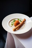 Un piatto bianco con un dessert composto da un boccone rotondo impanato, una fetta di pane e una salsa cremosa, guarnito con foglie verdi.
