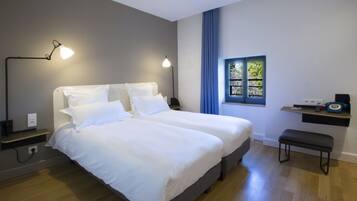 A modern bedroom with two beds, bedside lamps, a window with blue curtains, and a small bench.