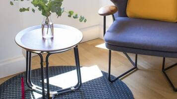 A room with a window, a chair, a small table with a plant, and a mat on the floor.