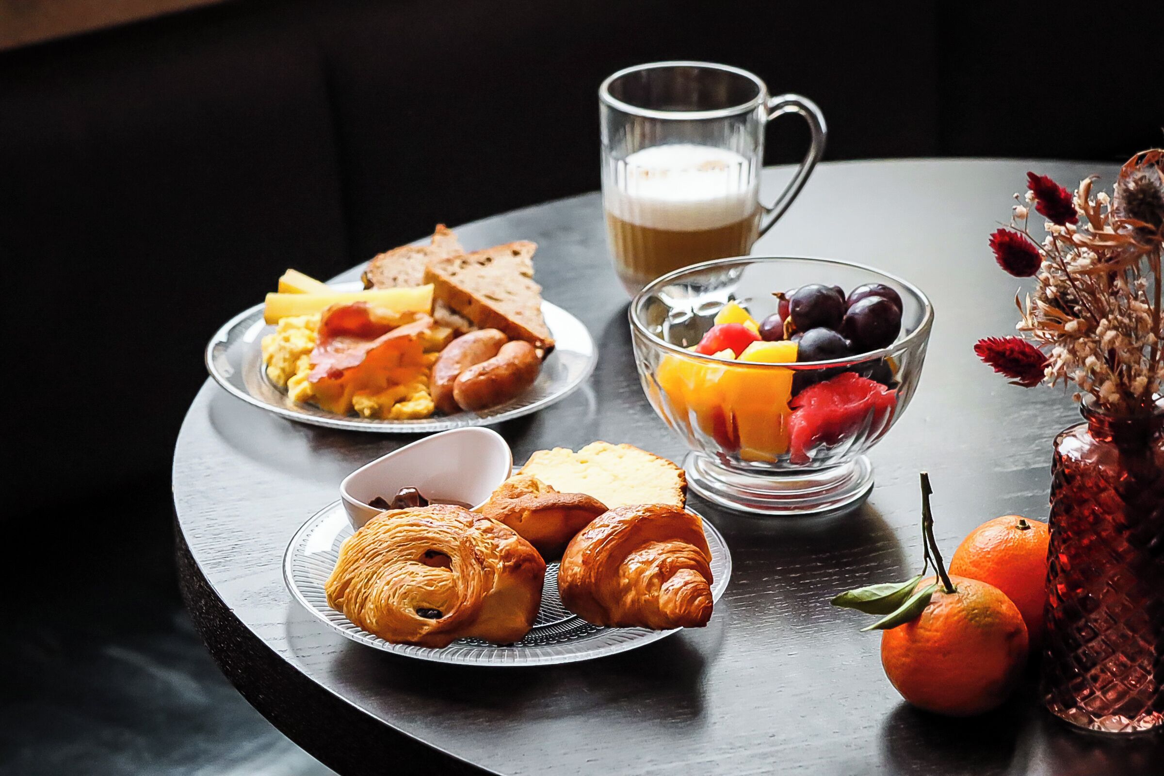 Un desayuno con cruasanes, tostadas, huevos y un bol de fruta sobre una mesa.