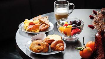 Un desayuno con cruasanes, tostadas, huevos y un bol de fruta sobre una mesa.