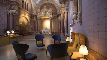 A spacious lobby with ornate architecture, including a high ceiling and detailed wall decorations. There are several seating areas with modern chairs and a sofa.