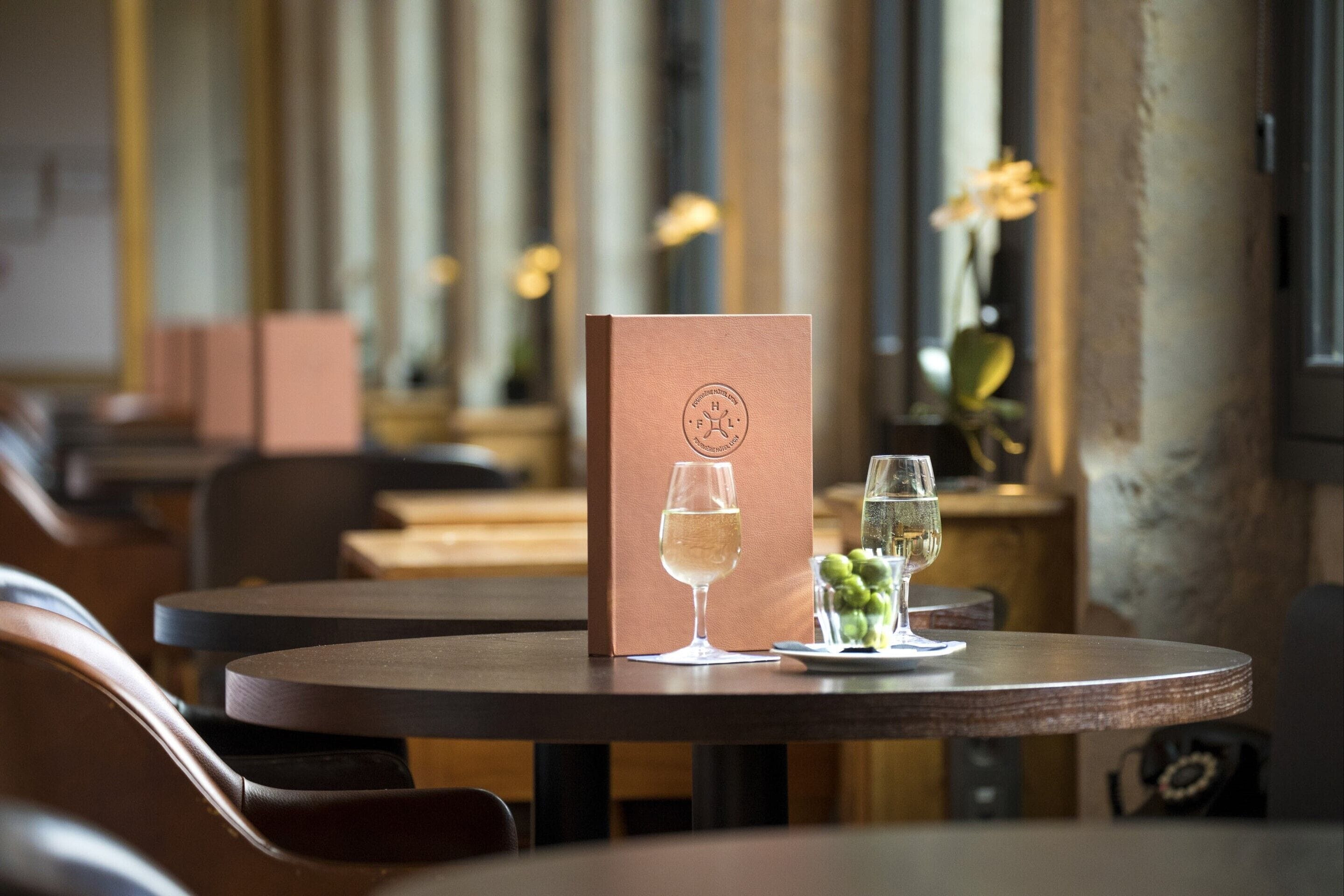 Una mesa de restaurante con un menú, dos copas de vino blanco y un florero con uvas verdes.