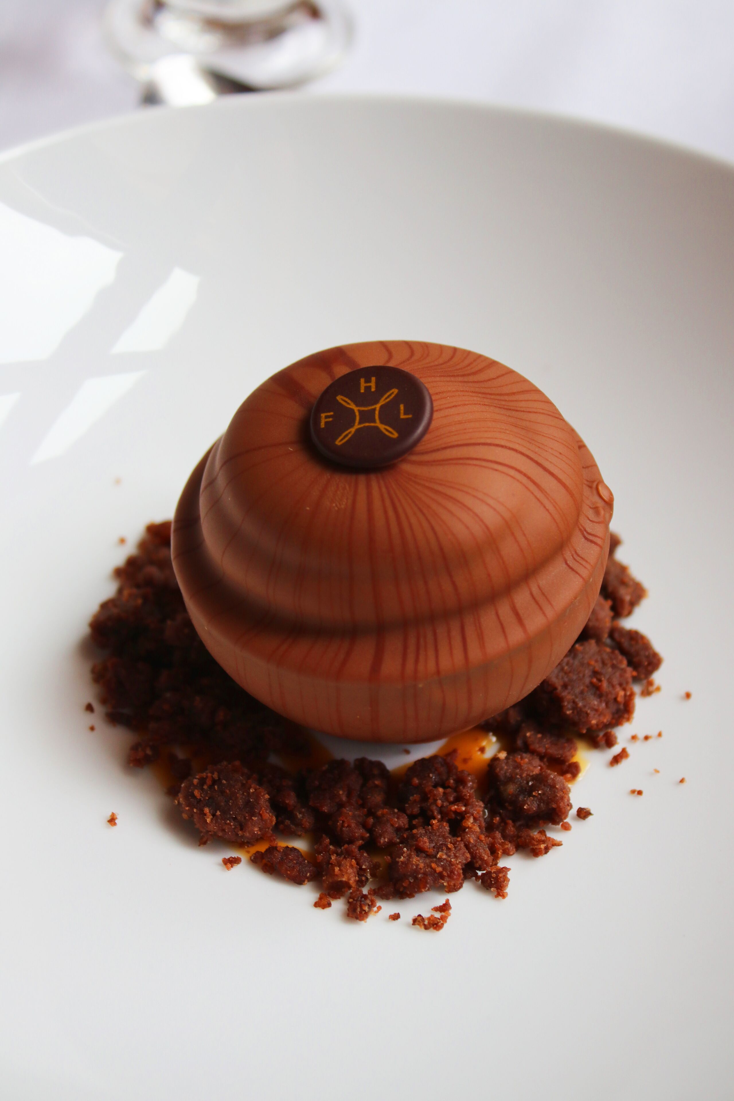 A dessert on a white plate with a chocolate dome and a chocolate piece on top.