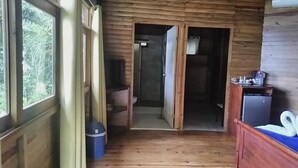 Een houten hutje met een houten vloer, een bed met een blauwe matras, een klein tafeltje met een waterkoker, en een raam met gordijnen.