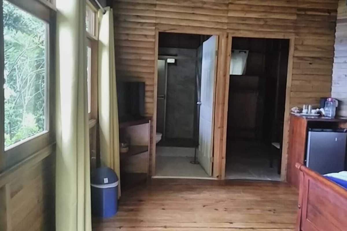 Een houten hutje met een houten vloer, een bed met een blauwe matras, een klein tafeltje met een waterkoker, en een raam met gordijnen.