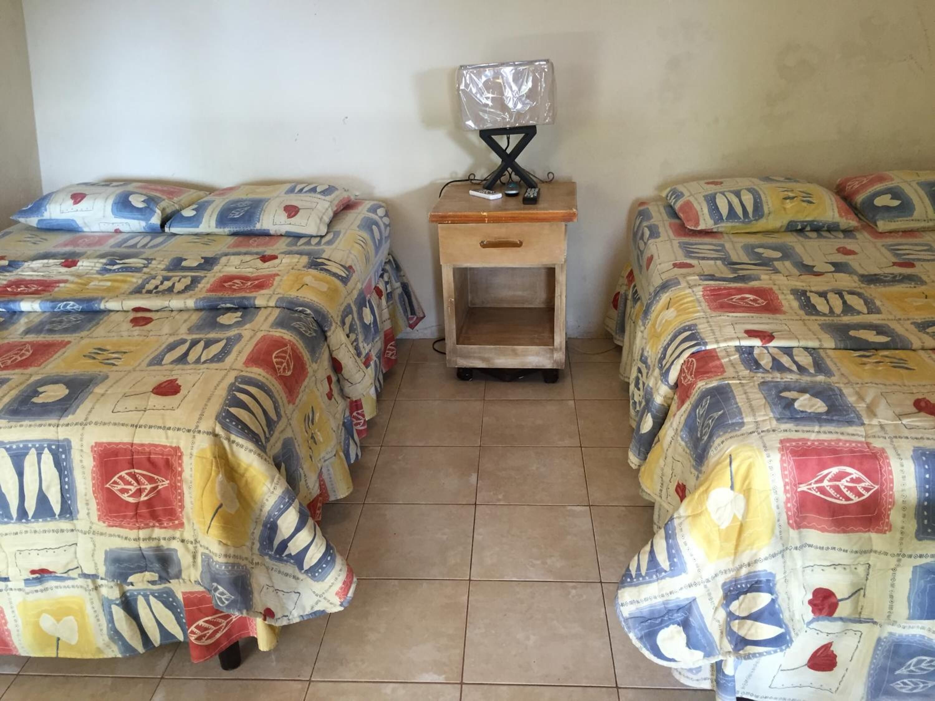 A room with two beds, each with a colorful quilt. There is a small wooden nightstand with a lamp between the beds.