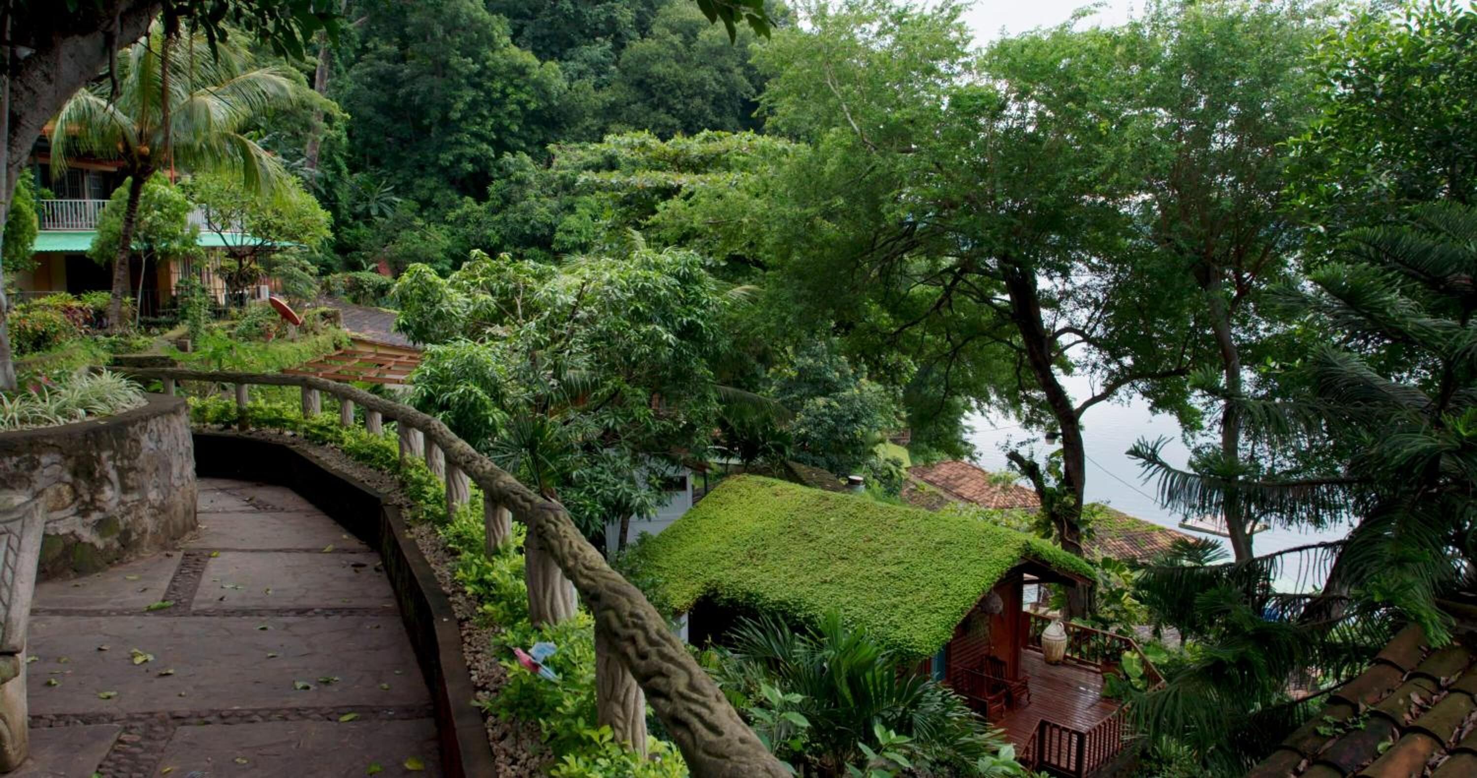 A lush garden with a wooden bridge, a small house with a thatched roof, and a view of a body of water.