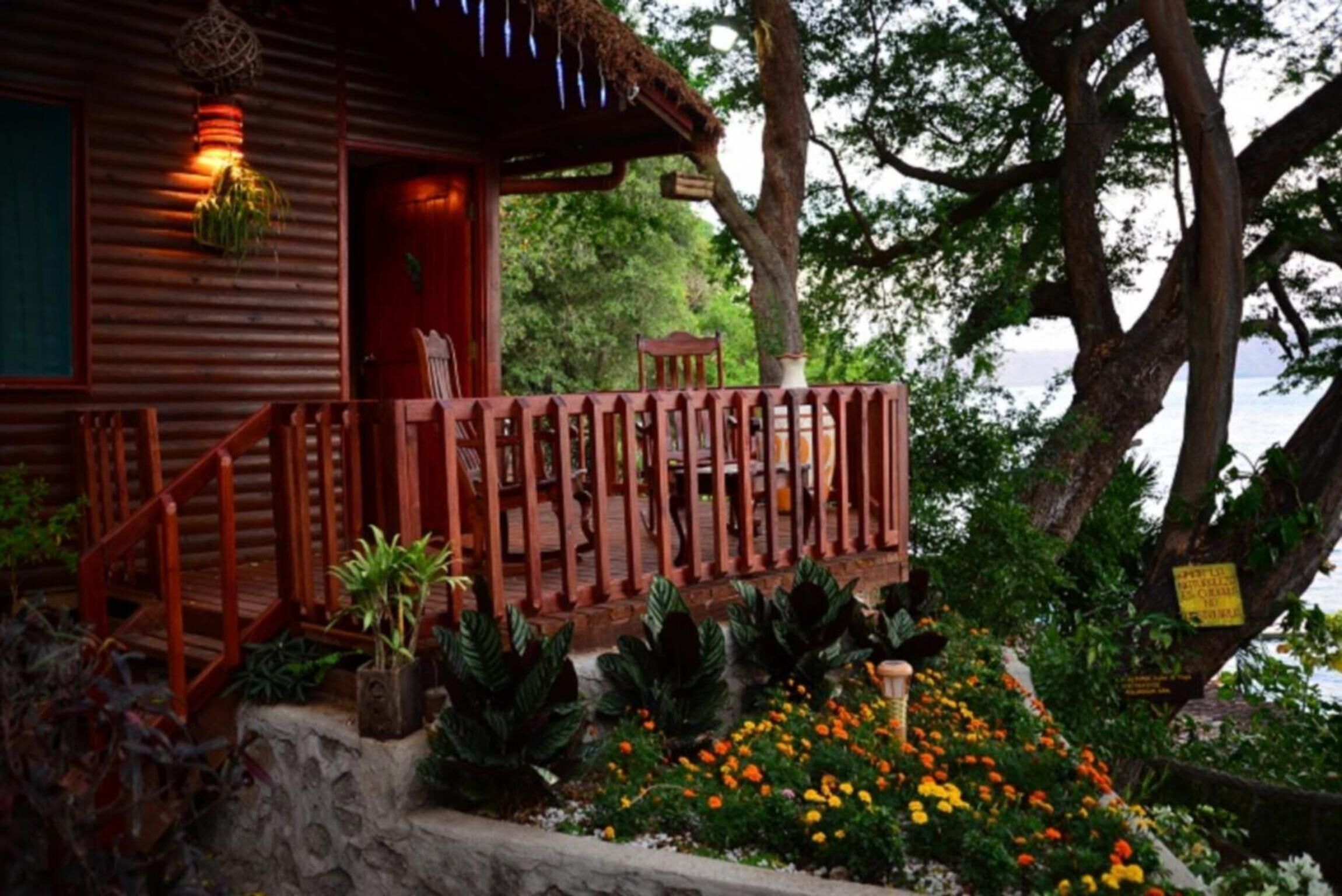 Una cabaña de madera con un porche de madera, rodeada de un jardín con diversas plantas y flores.