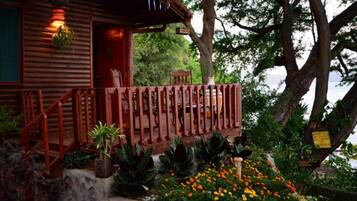 Una cabaña de madera con un porche de madera, rodeada de un jardín con diversas plantas y flores.